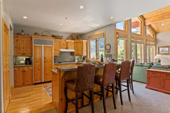The kitchen of Trails End Lodge 18 features wood cabinets, stainless steel appliances, and a breakfast bar with seating for three. The living area is visible in the background with a view of the mountains through large windows.