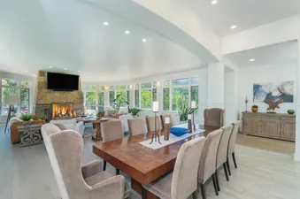 A spacious open-concept living and dining area in Turkey Creek with a stone fireplace, large windows, a wooden dining table, and upholstered chairs.