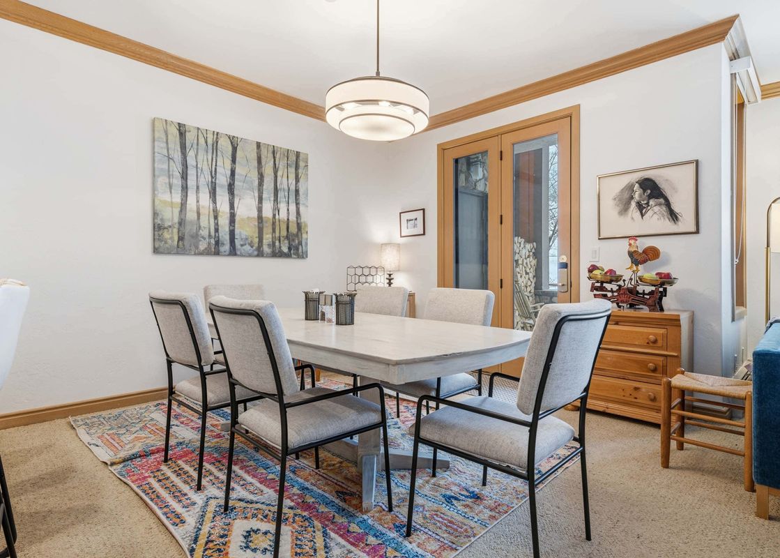 A rustic dining table with six chairs sits on a colorful rug in the dining area, illuminated by a modern chandelier, with a nature painting on the wall and French doors to the exterior.