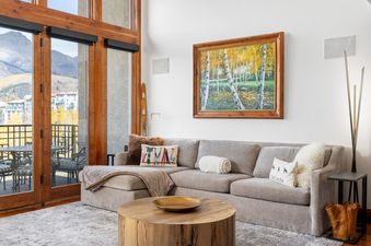 A bright living room with an L-shaped sofa, a wooden coffee table, and large windows offering an autumn mountain view.
