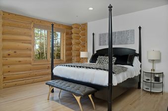 A rustic yet modern bedroom with a black four-poster bed, log walls, and a window overlooking trees.