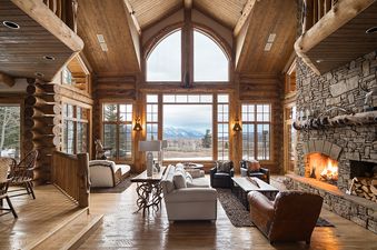 A luxurious and spacious rustic living room with a large stone fireplace, comfortable seating, and an expansive window view of snow-capped mountains.