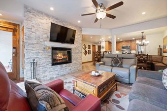 A cozy living room in The Charter F2505 with a stone fireplace, flat-screen TV, comfortable seating, and an open-concept view of the kitchen and dining area.