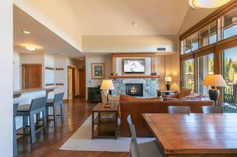A spacious living room with a stone fireplace and large windows overlooking a forest.
