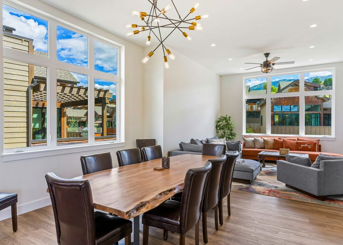 A modern living room with a large window looking out at a mountain view. The room has a large sofa, a coffee table, and a dining table with leather chairs.