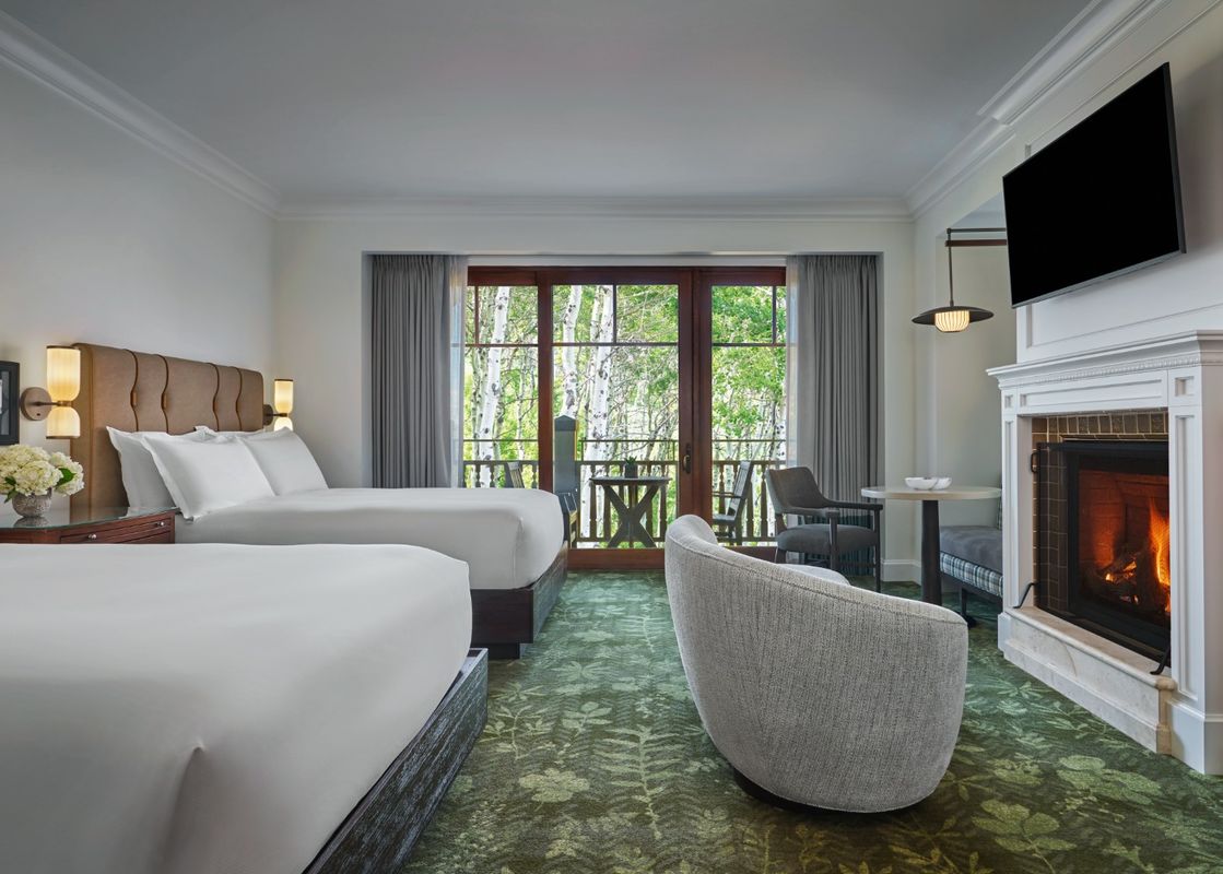 Two queen beds with white linens and brown headboards flank a bedside table with a vase of white flowers, while a fireplace with a lit fire and a mounted television are visible to the right, and a balcony overlooking a forest is straight ahead.