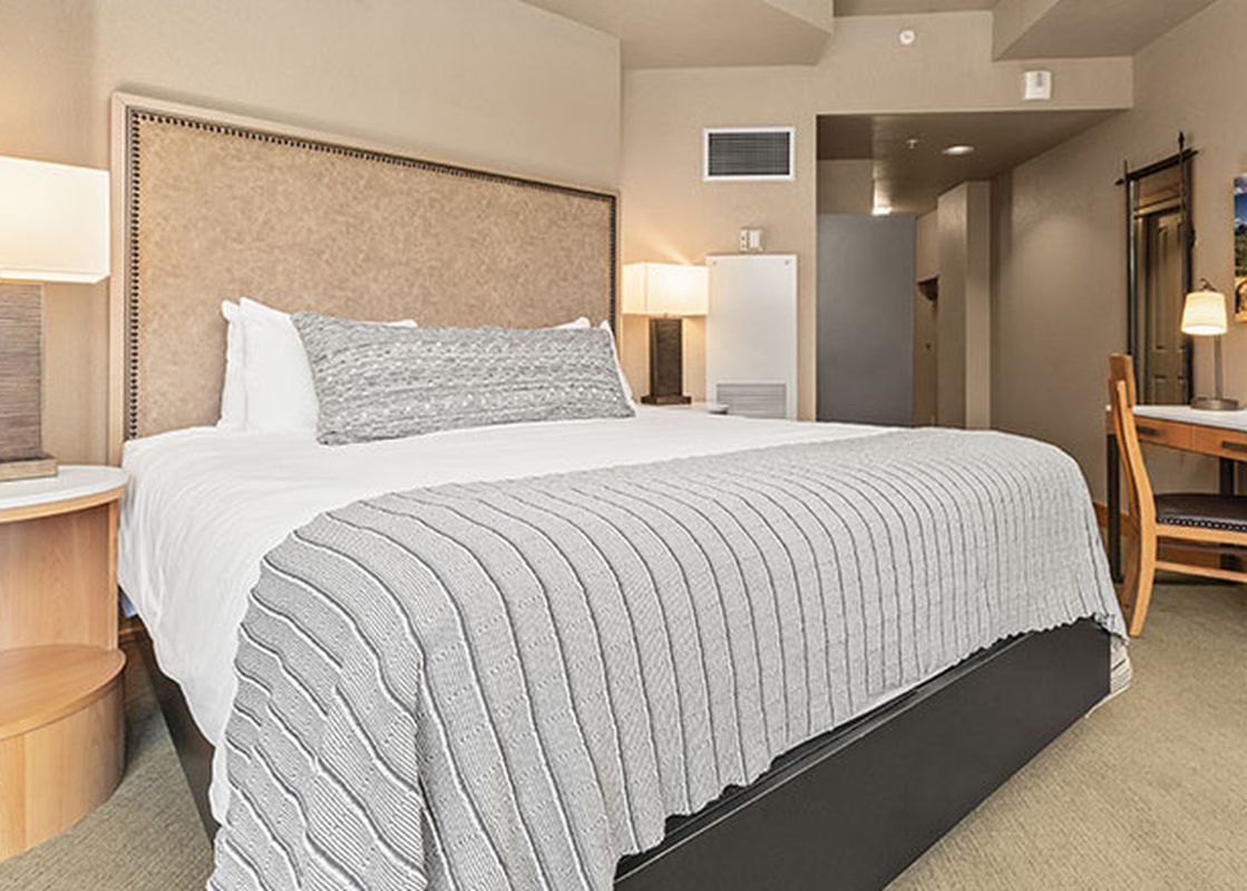 The image shows the Steamboat Grand Deluxe King, a spacious and inviting bedroom with a king-size bed, a bedside table with a lamp, and a desk area.