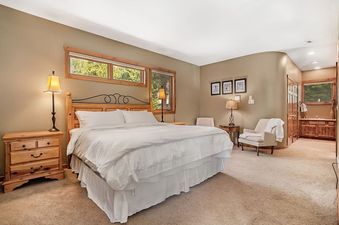 A spacious bedroom features a large bed with white linens, wooden furniture, and broad windows offering a view of lush greenery.
