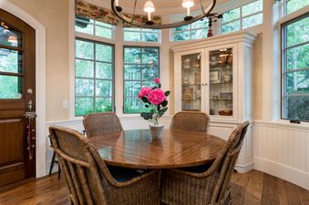 A dining table with four wicker chairs sits in a room with large windows and a white cabinet with glass doors. There are pink flowers in a vase in the middle of the table.