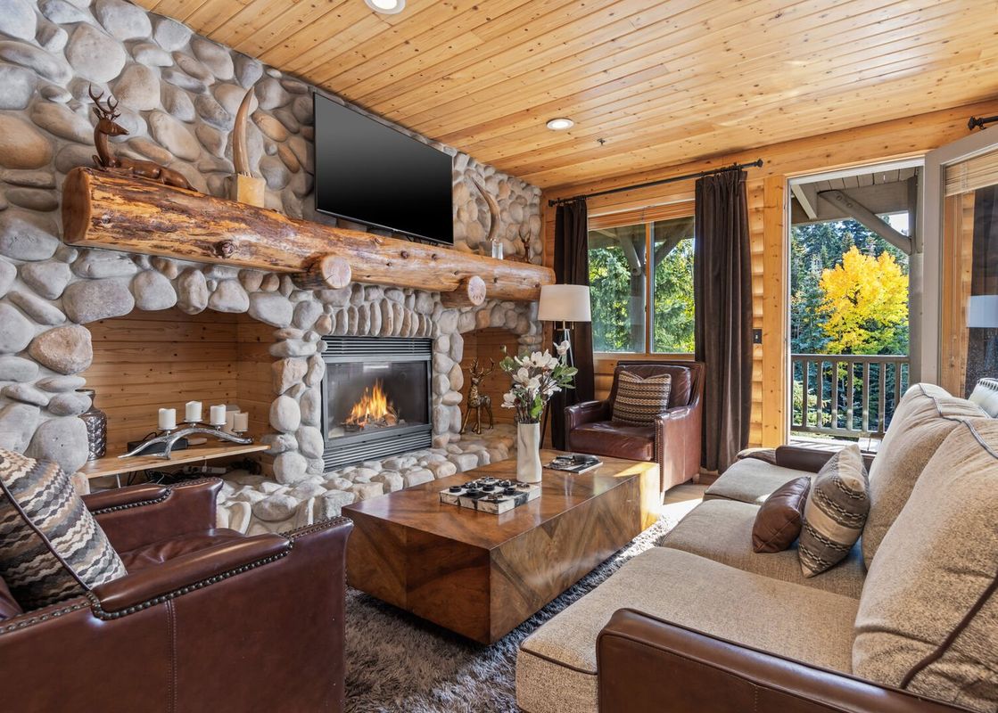 A rustic living room with a stone fireplace, comfortable seating, and a view of autumn foliage from the balcony.