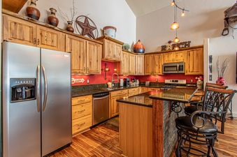 A kitchen with wooden cabinets, stainless steel appliances, and a granite countertop. There is a breakfast bar with two stools and a red accent wall.
