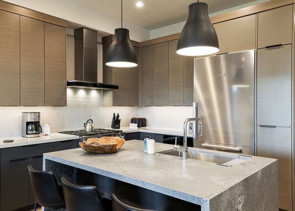 A modern kitchen with wood-grain cabinetry, gray countertops, stainless steel appliances, and two black pendant lights over an island with a sink.
