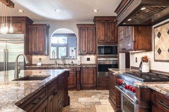 A spacious kitchen featuring dark wood cabinetry, granite countertops, stainless steel appliances, and a central island with a sink, under-cabinet lighting, and a window with an outdoor view.