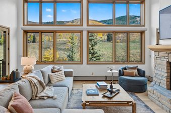 A cozy living room at Chalet Bravura features a large sectional sofa, a rustic coffee table, a stone fireplace with a TV mounted above it, and expansive windows showcasing a vibrant autumn landscape.