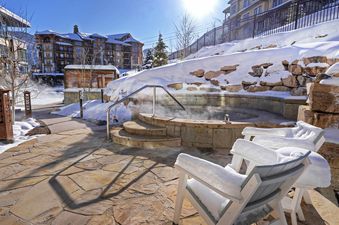 An outdoor hot tub in a snowy resort setting with snow covered chairs and a stone path.