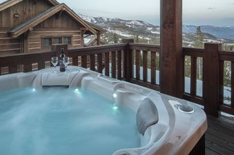 A private hot tub on the wooden balcony of Wolf Pack Hideaway, featuring a bottle of wine and two glasses, overlooks a snow-covered mountain landscape.