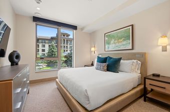 A modern bedroom at Assay Hill Lodge Unit 264 features a queen bed with teal pillows, a large window overlooking a resort, and contemporary furnishings, exuding a tranquil and inviting atmosphere.