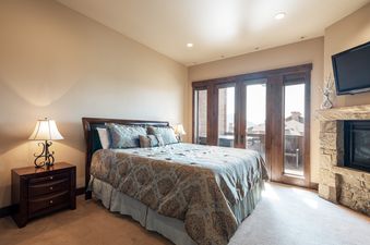 A master bedroom at 103 Juniper Landing with a large bed, nightstand, lamp, French doors, and a stone fireplace with a television.