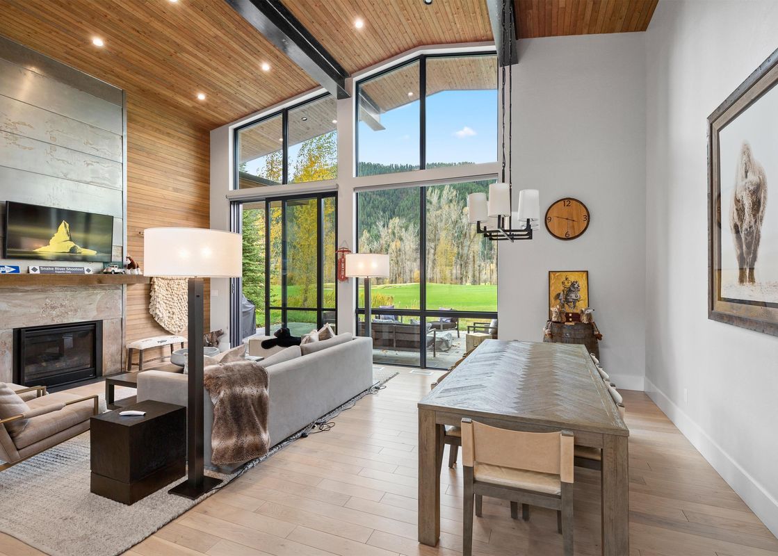 The living and dining area of the Fairway Cabin Two Bedroom with Golf View features an open layout with high ceilings, large windows overlooking a golf course and mountains, a fireplace, and a wooden dining table.