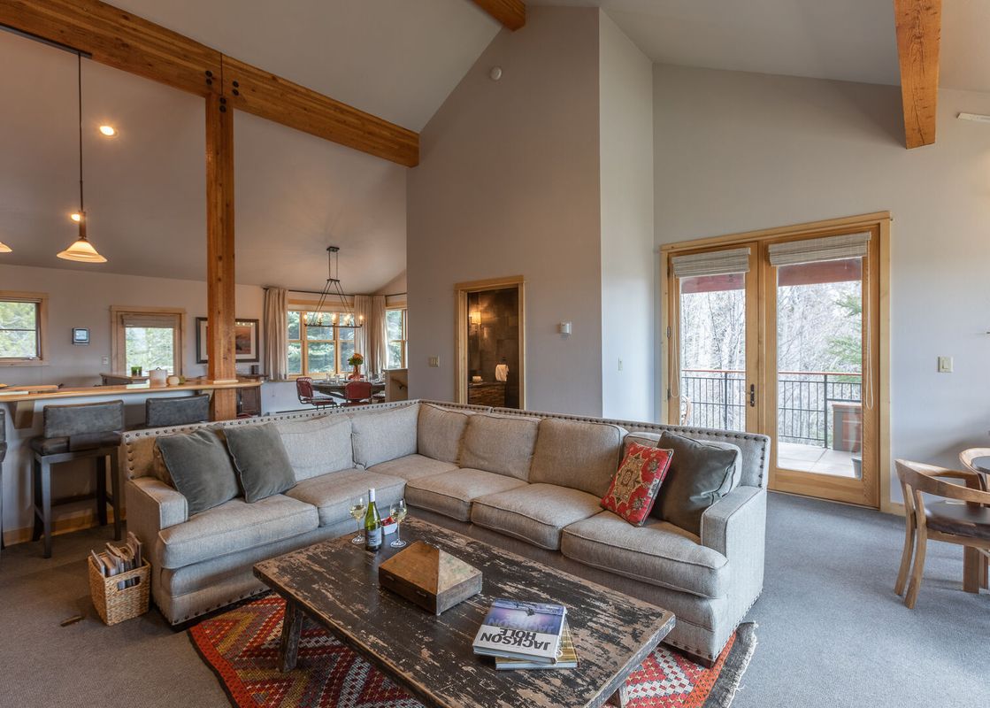 A cozy, open-plan living room with a large sectional sofa, rustic coffee table, vaulted ceiling with wood beams, and glass doors leading to a balcony.