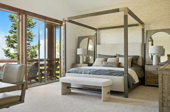 A king-sized four-poster bed with neutral upholstered headboard and grey and beige linens, flanked by bedside tables with lamps and mirrors, sits opposite a private balcony with nature views.