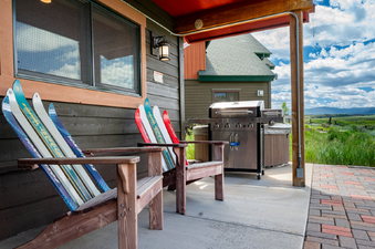 An inviting outdoor patio at 2002 Lupine Lane features two Adirondack chairs with ski backs, a large BBQ grill, and a hot tub, all set against a backdrop of rolling green hills under a bright, cloudy sky.