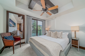 The bedroom in Lumiere Two Bedroom Residence features a king bed with plush bedding and a cozy throw blanket, a ceiling fan, large windows with views of the trees, and a bedside table with a lamp.