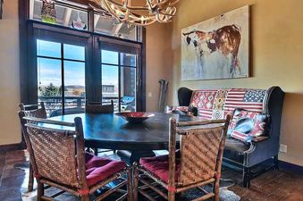 A rustic dining area with a round wooden table, chairs with woven backs and red cushions, and a flag-patterned bench below a cow painting, featuring a window with an exterior view.