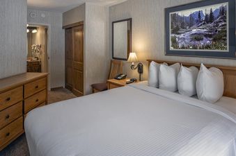 A cozy bedroom in the Building 4 One Bedroom Beaver Run Resort with a queen-sized bed, a nightstand with a lamp, a wooden dresser, and a large landscape painting above the bed.
