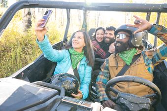 A group of four friends takes a selfie while riding an ATV through a sunny, autumn forest.