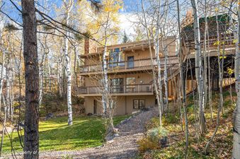 A tan wooden house with multiple decks and a large stone fireplace sits nestled in the forest with trees surrounding it.
