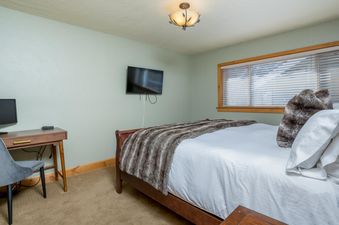 The serene bedroom at Krystal Villa 5 features a bed with white linens and a faux fur throw, a wall-mounted TV, a wooden desk with a chair, and a window with blinds, all illuminated by a ceiling light.