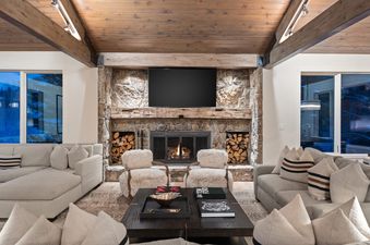 A cozy, luxurious great room at the Casa de Gondola featuring a large stone fireplace with a television, two plush armchairs, and two comfortable sectionals, all under a vaulted wooden ceiling.