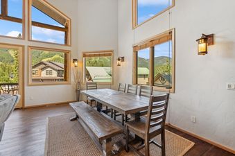 A rustic dining area with a long wooden table, a bench, and chairs, with large windows overlooking a mountainous landscape and other homes.