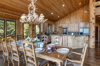 The American Flag Solamere dining and kitchen area features wooden interiors, an antler chandelier, a set dining table, and a well-appointed kitchen with modern appliances and a large window.