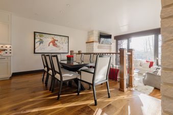 A modern dining room with a dark wood table, light chairs, and a large ski-themed painting on the wall, open to a sunlit living area with a view of snow-covered trees.