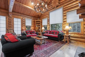 The rustic living room at Obsidian Jackson Hole features log walls, a vaulted wooden ceiling, and a large antler chandelier illuminating two black sofas and an armchair with red pillows, centered around a wooden coffee table and a patterned rug.