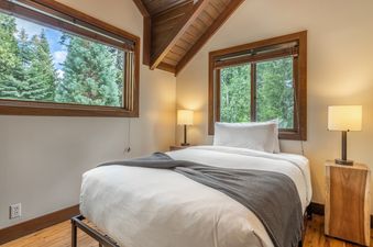 A cozy bedroom in Cedar Crest Owl Cottage 9 with a bed, two windows offering views of trees, and two bedside lamps.