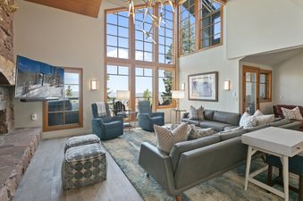 A spacious and bright living room in Stag Lodge 10 featuring tall windows with outdoor views, comfortable seating, a fireplace, and a large television.