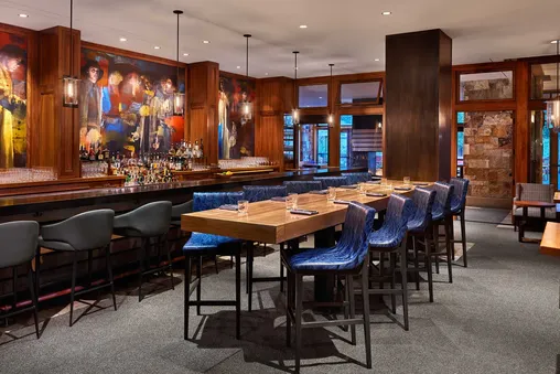 A modern bar with dark wood paneling and artwork, featuring a long table with blue upholstered chairs and a bar with black stools.