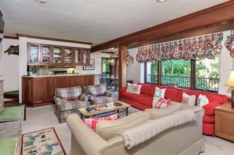 The living area of Plaza Lodge 3 Bedroom Gold Unit 2 showcases comfortable seating with two plaid armchairs, a red sectional sofa, and a cream-colored sofa arranged around a coffee table, with a built-in wet bar featuring wooden cabinetry on the left and