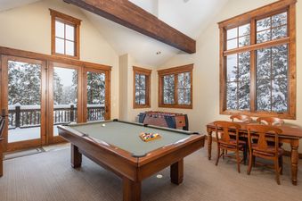A spacious game room at Last Nickel Lodge with a pool table, foosball table, and a dining area, all bathed in natural light from large windows offering a snowy outdoor view.