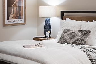 A cozy bedroom with a wooden bed, white linens, a bedside table with a lamp, and a framed Western poster on the wall.