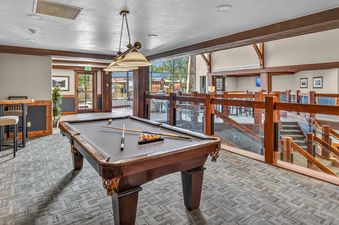 A pool table with billiard balls and cues is the focal point of a recreational room at Crystal Peak Lodge 7407, featuring wooden beams, geometric carpet, and large windows with outdoor views.