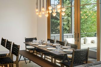 A modern dining room features a large dark wood table set with white plates and glassware, accompanied by black woven chairs and a long bench, all illuminated by a cluster of pendant lights hanging overhead.