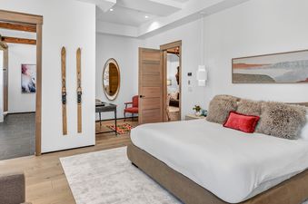 A modern and cozy master bedroom featuring a large bed with a brown upholstered headboard, white bedding, and decorative pillows. The room includes wooden accents, modern lighting, and artwork, with a glimpse into an adjoining bathroom.