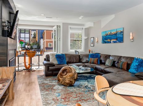 A modern living room featuring a brown leather sectional sofa with colorful pillows, a unique wooden coffee table, a breakfast bar, and large bi-fold doors opening to an exterior view.