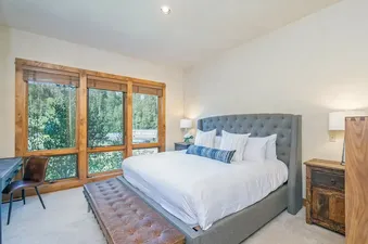 A beautifully furnished bedroom at Sunshine at the terraces with a grey tufted bed and large windows displaying a forest view.