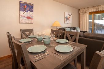 The dining area in Silverado I C101 features a wooden table set with light blue plates and glasses, complemented by a rustic lamp and a vintage skiing poster on the wall, with a comfortable living area visible in the background.