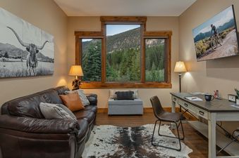 A cozy, sunlit living area boasts a luxurious dark leather sofa adorned with plush cushions, facing a wall-mounted flat-screen TV and a functional wooden desk, all complemented by a chic faux cowhide rug on rich hardwood floors.